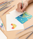 Hand using Capped Iridescent Glitter Gel Ink Pen Set Of 12 to create colorful glitter art on postcard with scattered pens nearby
