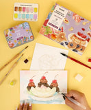 Hand painting a dessert watercolor scene with brushes and paints from Grabie Quarterly Watercolor Coloring Club set